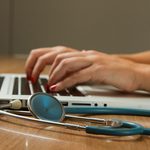 Dłonie piszące na laptopie, obok na drewnianym biurku leży stetoskop