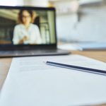 Close up of pencil and sheet of paper on the table. Laptop screen with excited female teacher communicating during video call in the background